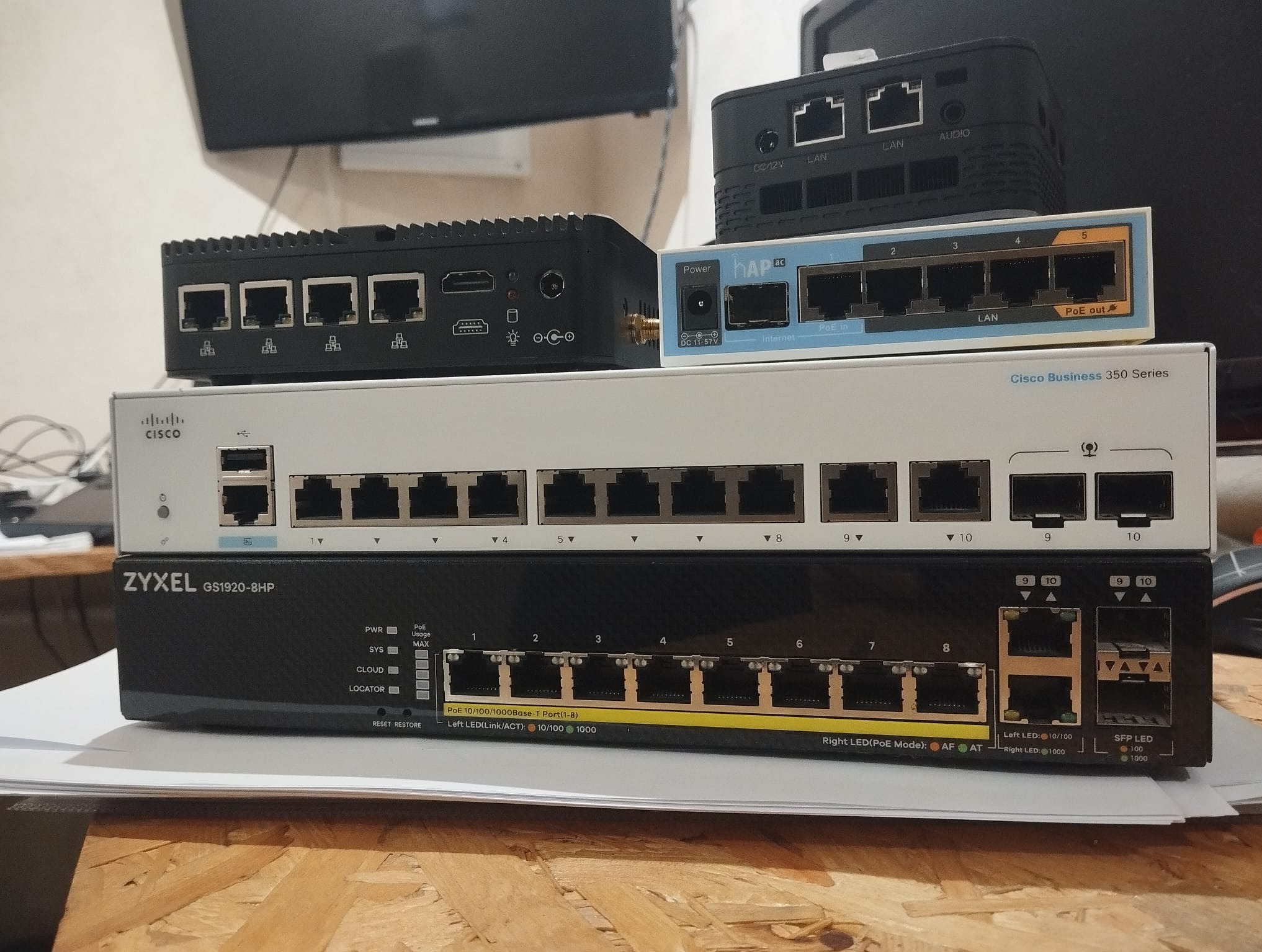 Stos routerów i switchy sieciowe różnych marek (Cisco, Zyxel, MikroTik) ułożone jeden na drugim na drewnianym blacie. Widoczne porty Ethernet i oznaczenia modeli.