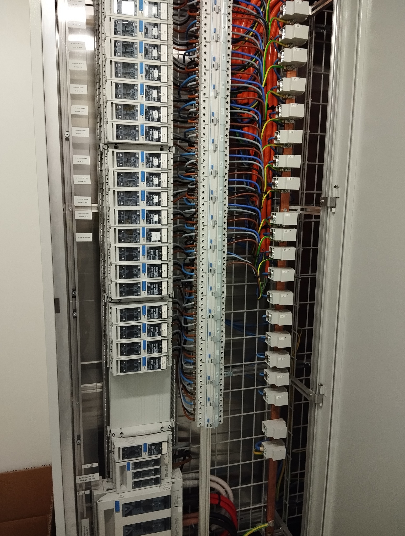 Rozdzielnica elektryczna z modułami, przewodami i listwami montażowymi w metalowej obudowie. Widoczne oznaczenia na modułach i przewodach. Ujęcie z bliska, oświetlenie neutralne.