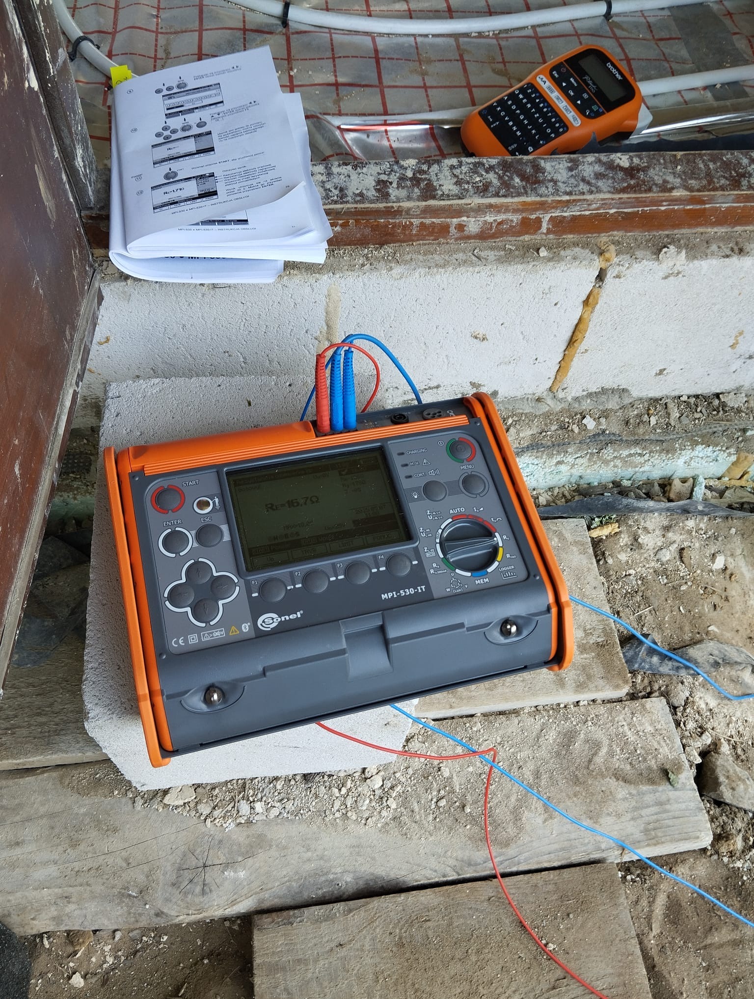 Miernik elektryczny Sonel MPI-530-IT na budowie, wyświetlający wynik pomiaru. Widoczne kable pomiarowe, instrukcja obsługi i etykieciarka Brother.