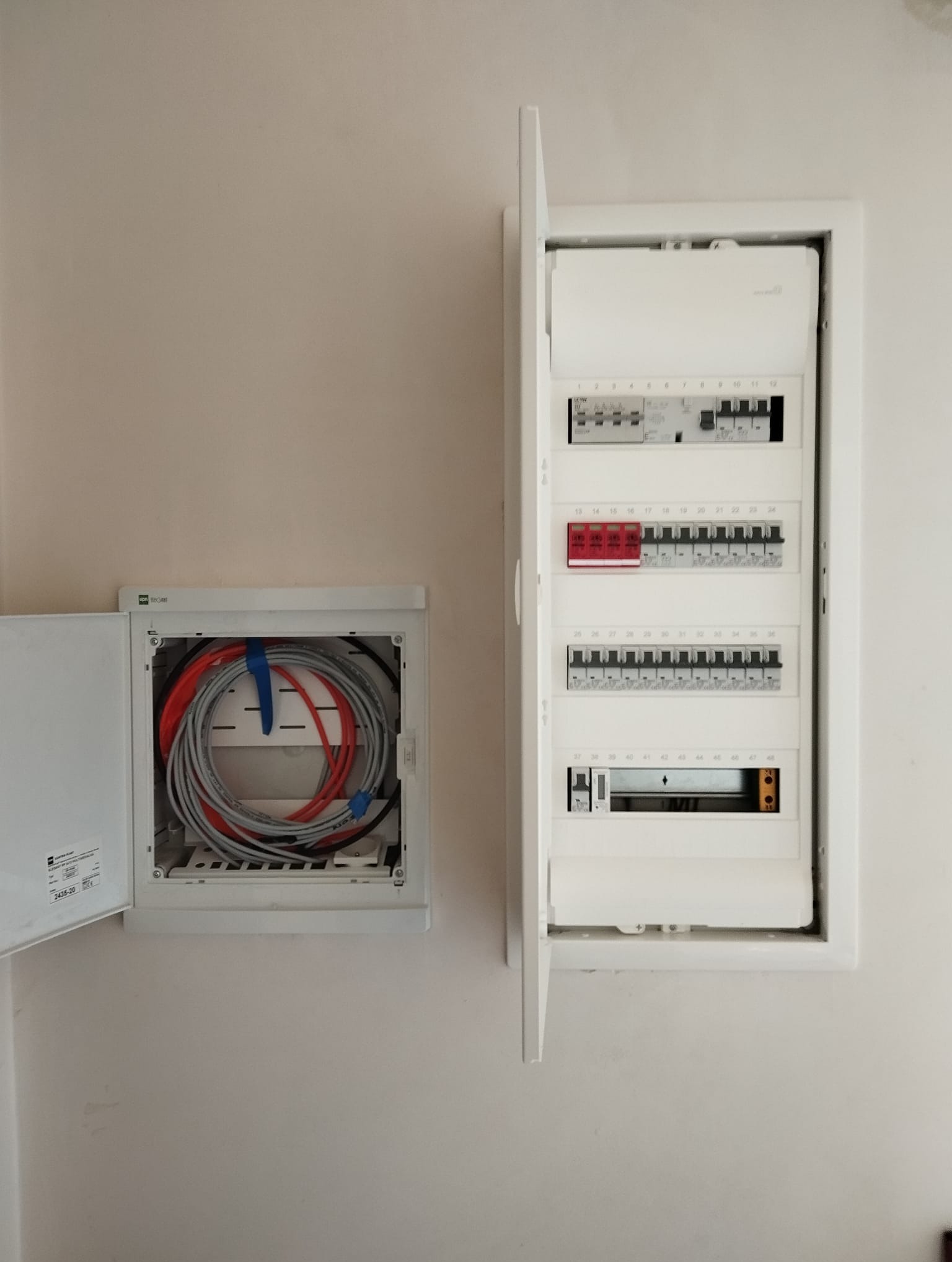 Otwarta skrzynka elektryczna z bezpiecznikami i przewodami w ścianie. Widoczne kable ułożone w pętle w drugiej skrzynce. Białe elementy na jasnym tle ściany.