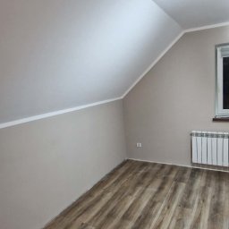 Łukasz Hanas - Pokój na poddaszu po remoncie: świeżo pomalowane ściany w beżowym odcieniu, panele podłogowe imitujące drewno, biały grzejnik pod oknem. Skos dachu.