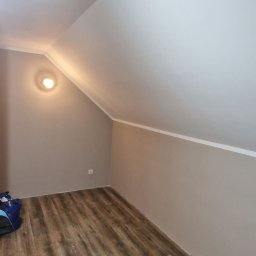 Łukasz Hanas - Pokój na poddaszu po remoncie: drewnopodobna podłoga, beżowe ściany, biały sufit ze skosem i listwami. Widoczna lampa sufitowa i fragment worka budowlanego.