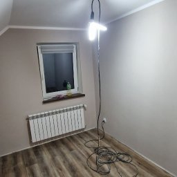 Łukasz Hanas - Pokój po remoncie: drewnopodobna podłoga, beżowe ściany, biały grzejnik pod oknem. Na suficie lampa robocza. Widok na poddasze.
