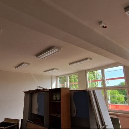 Łukasz - Jasne wnętrze z białymi ścianami i sufitami, oświetlone przez szereg lamp. Widoczne okna z widokiem na zieleń i budynki w oddali. Częściowo widoczne meble.