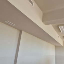 Łukasz - Jasne wnętrze po malowaniu: kremowe ściany i sufit z beżowymi akcentami, widoczne kratki wentylacyjne i lampy sufitowe. Minimalistyczny design, świeżo po remoncie.