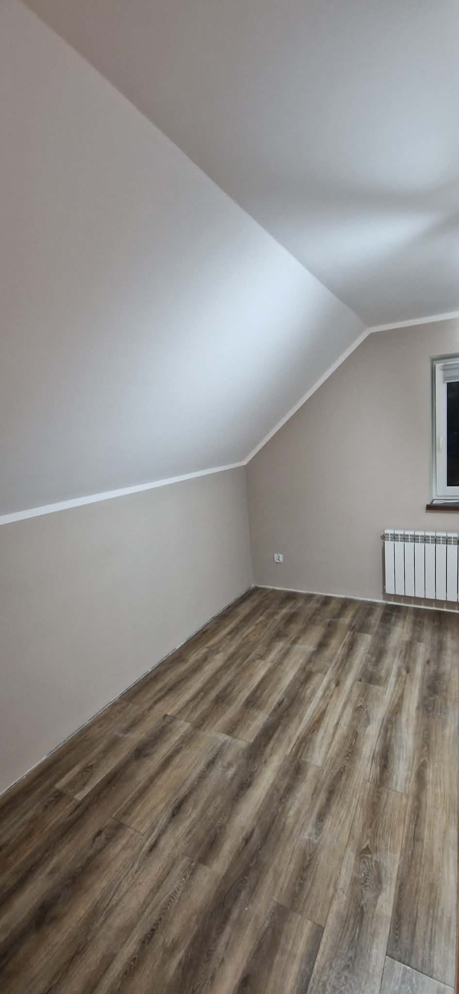 Pokój na poddaszu po remoncie: świeżo pomalowane ściany w beżowym odcieniu, panele podłogowe imitujące drewno, biały grzejnik pod oknem. Skos dachu.