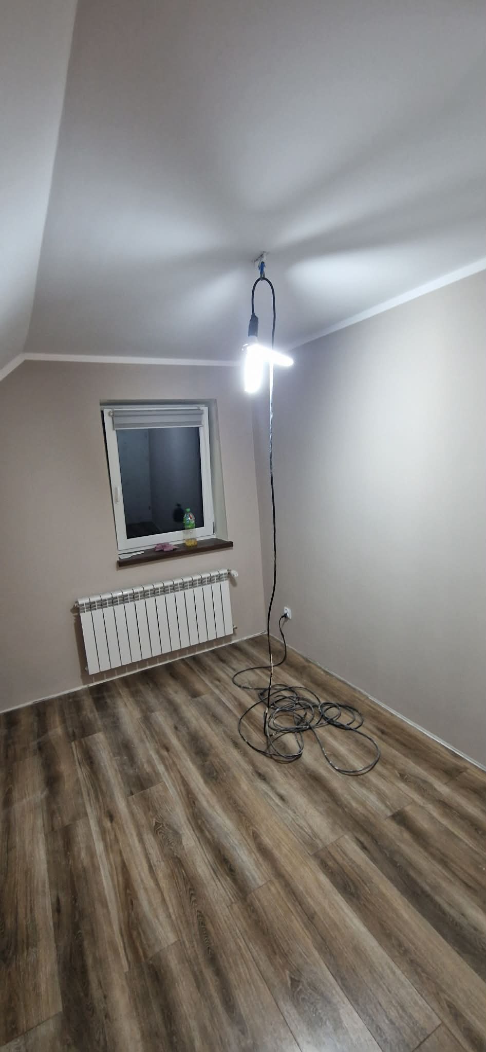 Pokój po remoncie: drewnopodobna podłoga, beżowe ściany, biały grzejnik pod oknem. Na suficie lampa robocza. Widok na poddasze.