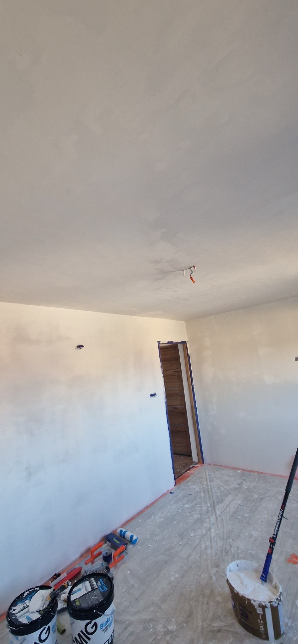 Wnętrze po szpachlowaniu: gładkie, białe ściany i sufit, zabezpieczone taśmą drzwi, narzędzia i pojemniki z masą szpachlową na podłodze. Widok na przygotowane pomieszczenie.