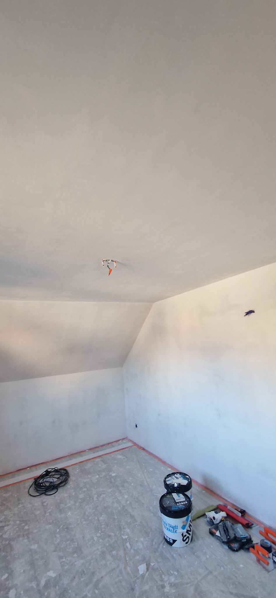 Poddasze po szpachlowaniu, przygotowane do malowania. Widoczne wiaderka z masą szpachlową, narzędzia i zabezpieczona folią podłoga. Ściany i sufit gładkie, białe.