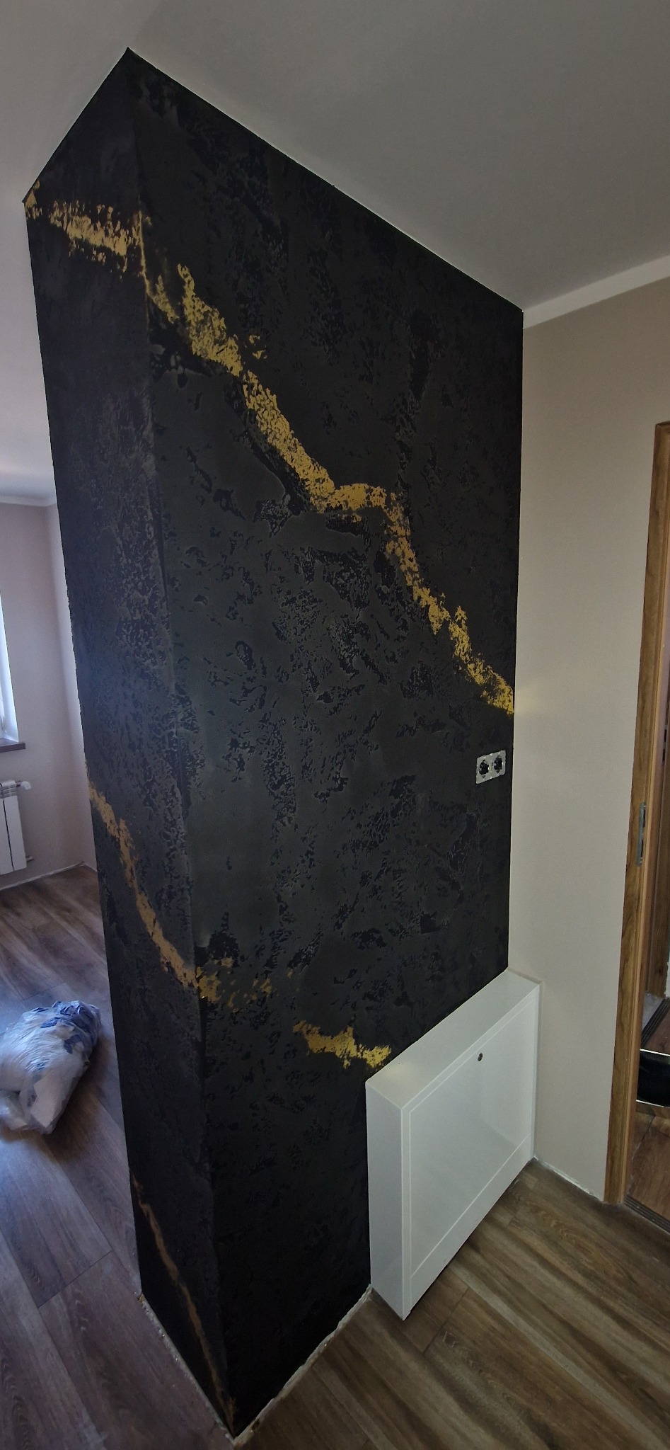 Ściana z dekoracyjną, czarno-złotą strukturą, imitującą marmur, w rogu pomieszczenia. Widoczny fragment grzejnika i podłogi z paneli.