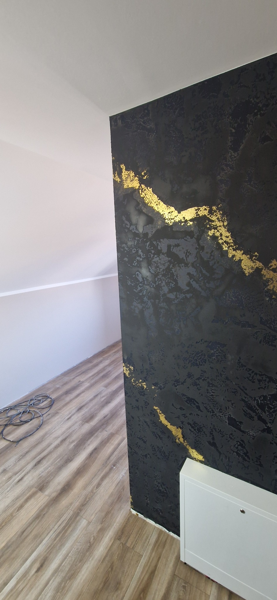 Ściana z dekoracyjną, czarno-złotą fakturą w nowocześnie wykończonym wnętrzu. Widoczna podłoga z paneli i fragment grzejnika. Unikalne wykończenie ściany.