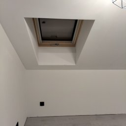 Marbud - Białe wnętrze poddasza z oknem dachowym w drewnianej ramie. Widoczne gładkie ściany, szara podłoga i fragment lampy. Minimalistyczny design.