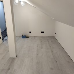 Marbud - Jasne panele podłogowe w pokoju na poddaszu z białymi ścianami i nowoczesną lampą sufitową. Widoczne gniazdka elektryczne i narzędzia w tle.