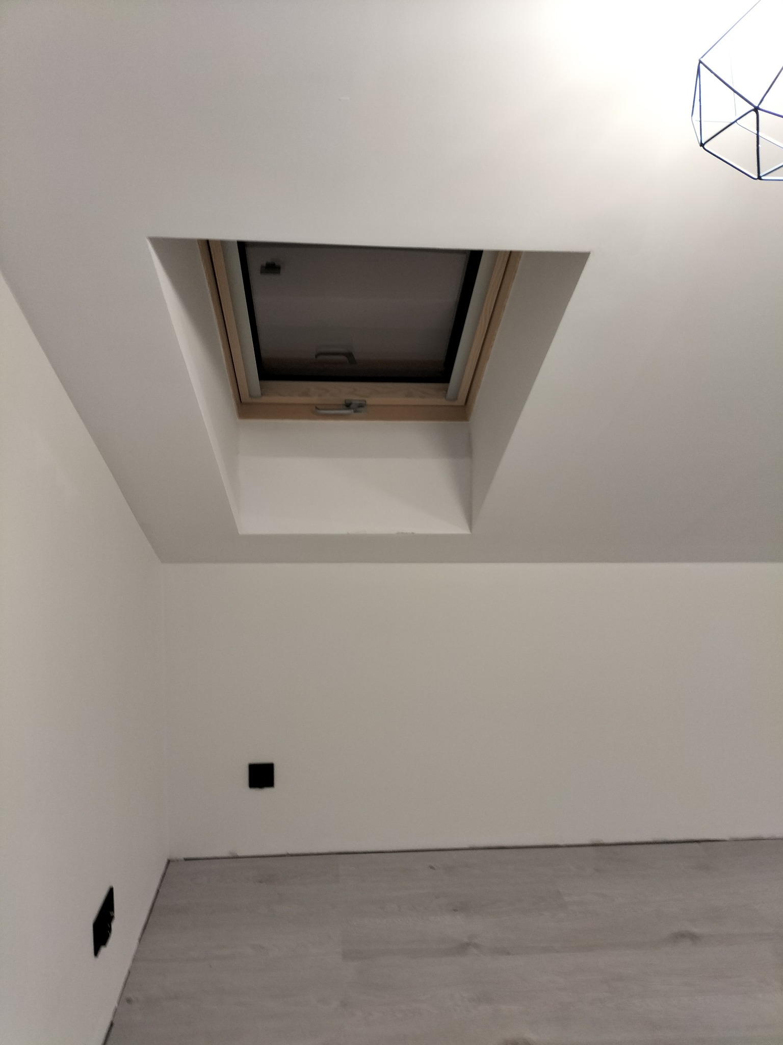 Białe wnętrze poddasza z oknem dachowym w drewnianej ramie. Widoczne gładkie ściany, szara podłoga i fragment lampy. Minimalistyczny design.