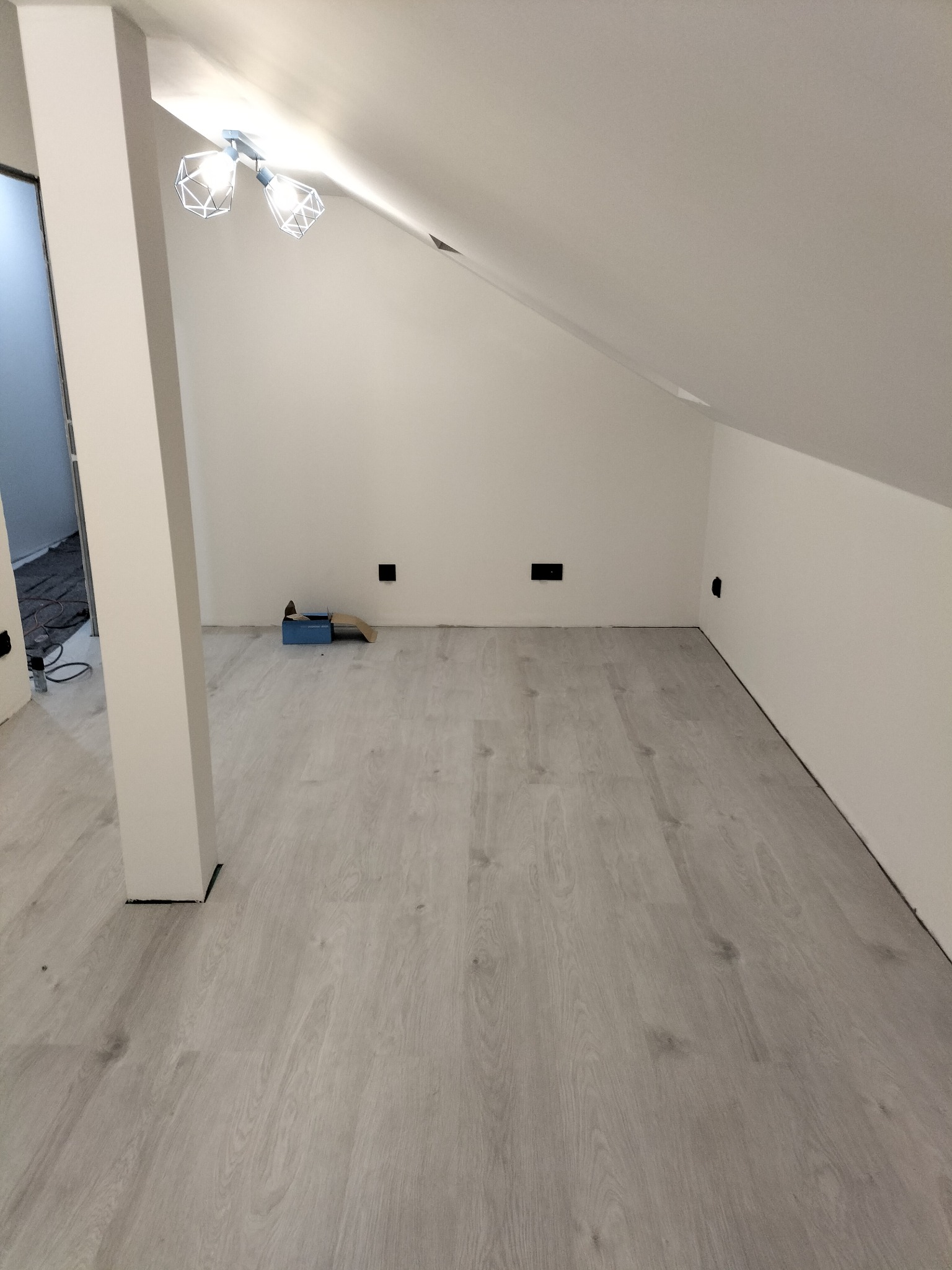 Jasne panele podłogowe w pokoju na poddaszu z białymi ścianami i nowoczesną lampą sufitową. Widoczne gniazdka elektryczne i narzędzia w tle.
