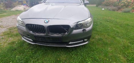 Uszkodzony przód szarego BMW F10 na trawie. Brak zderzaka, uszkodzona lampa i atrapa chłodnicy. Widoczne ślady naprawy blacharskiej. Ujęcie z bliska, na zewnątrz.