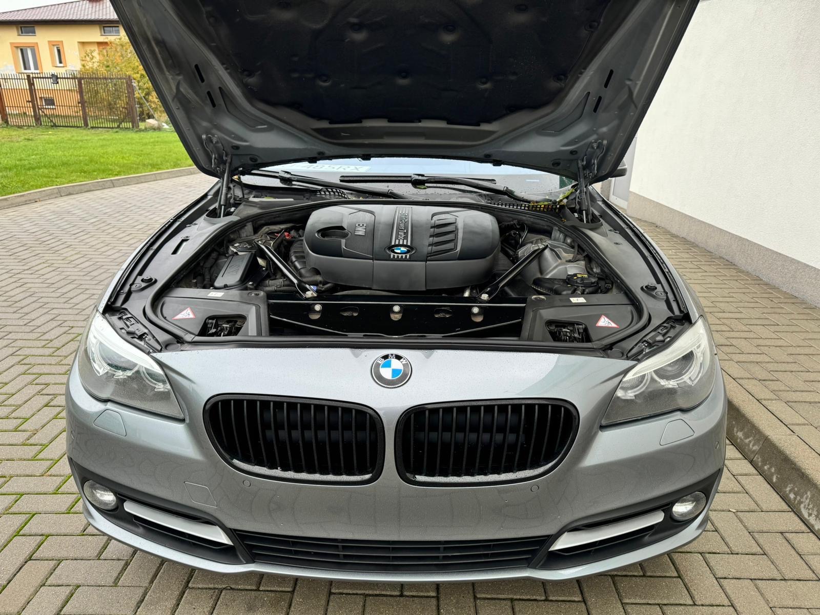 Szary samochód BMW z otwartą maską, widoczny silnik. Ujęcie z przodu, na pierwszym planie atrapa chłodnicy i reflektory. Tło stanowi budynek i fragment trawnika.