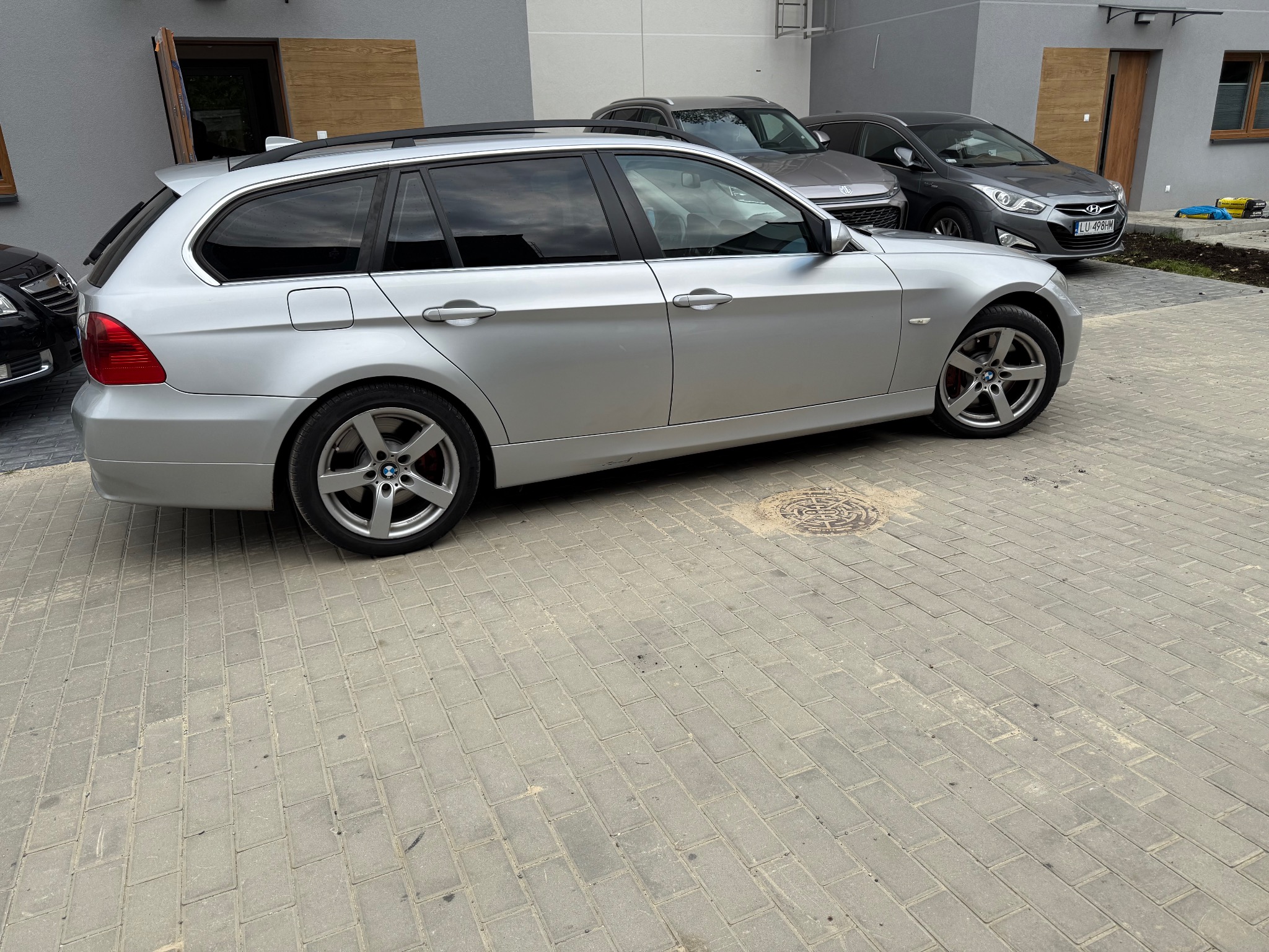 Srebrne kombi BMW serii 3 E91 na kostce brukowej, z widocznymi felgami aluminiowymi i innymi autami w tle. Ujęcie z boku, podkreślające sylwetkę pojazdu.