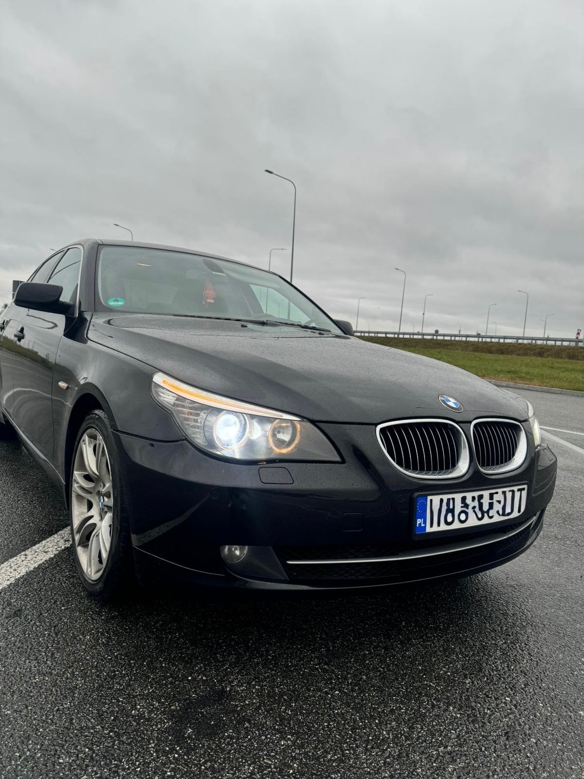 Czarny samochód BMW serii 5 E60 z włączonymi światłami mijania, zaparkowany na mokrym asfalcie w pochmurny dzień. Widoczny przód pojazdu i tablica rejestracyjna.