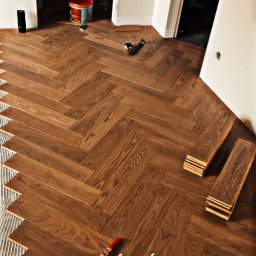 Przedsiębiorstwo Wielobranżowe Konrad Małkowski - Ukończona podłoga z parkietu ułożonego w jodełkę, widoczne narzędzia i klej. Częściowo widoczne drzwi i ściany. Ciepłe oświetlenie.