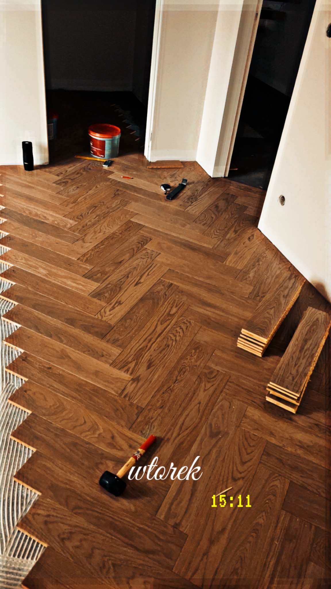 Ukończona podłoga z parkietu ułożonego w jodełkę, widoczne narzędzia i klej. Częściowo widoczne drzwi i ściany. Ciepłe oświetlenie.