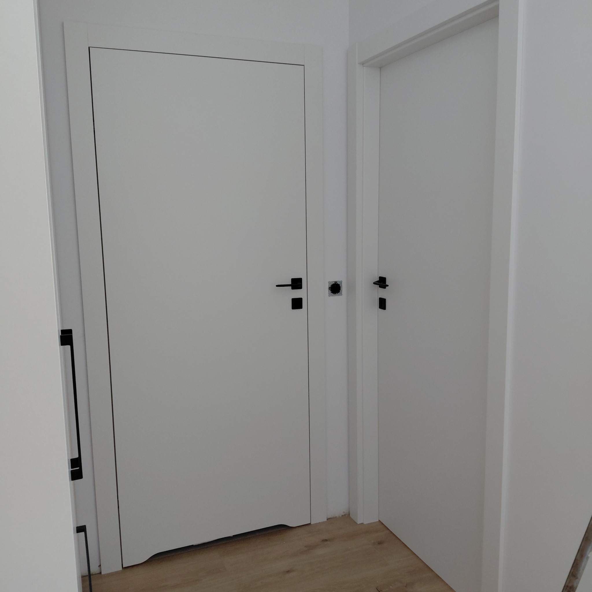 Dwa białe, zamontowane drzwi z czarnymi klamkami w minimalistycznym stylu. Jedne drzwi są zamknięte, drugie otwarte, ukazując fragment podłogi.