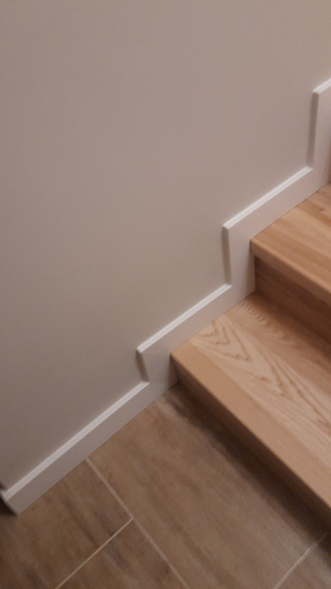 Wykończone drewniane schody z białą listwą przypodłogową i panelami podłogowymi w odcieniach beżu. Detal połączenia schodów ze ścianą.