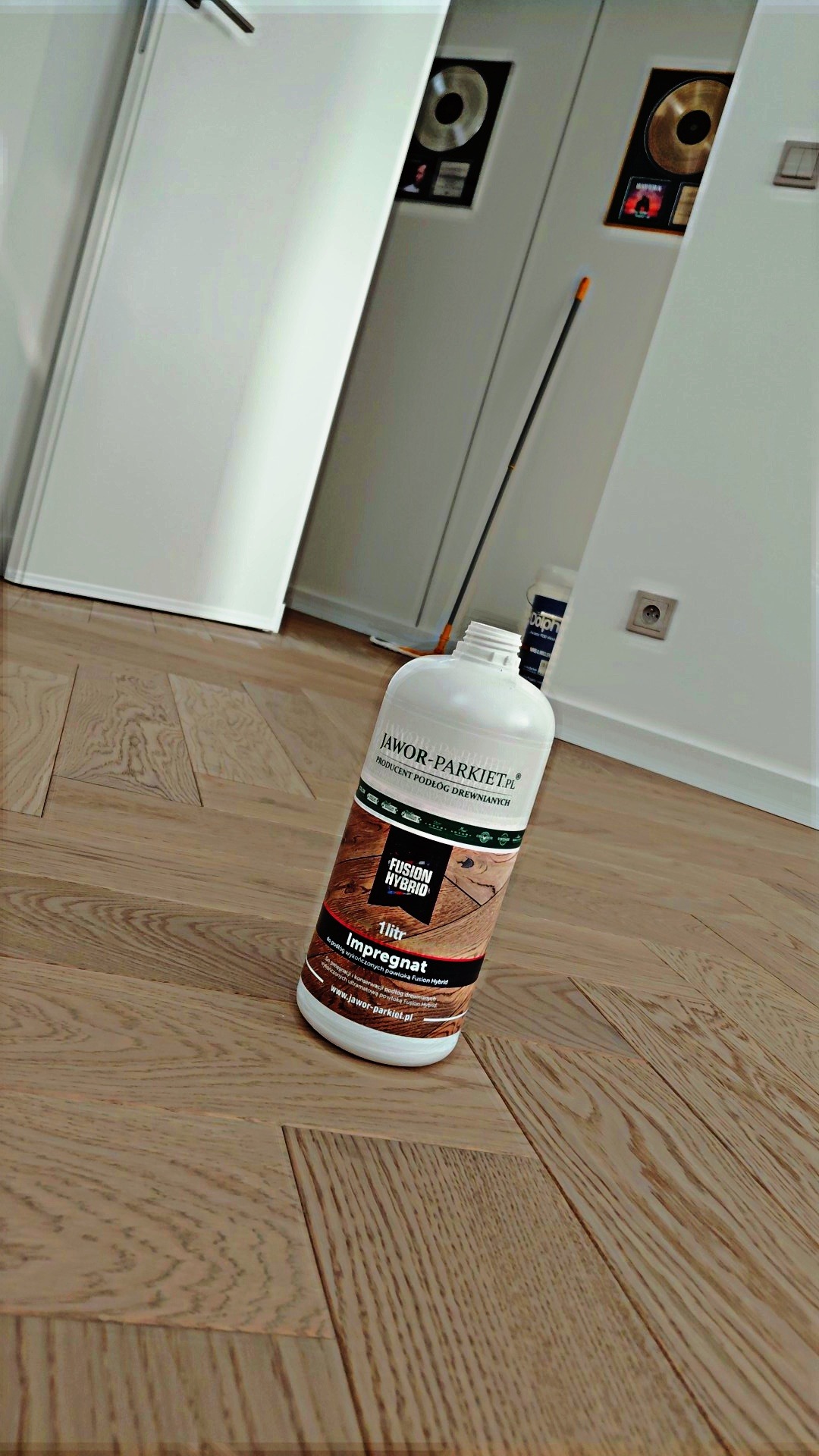Butelka impregnatu Jawor-Parkiet na parkiecie, w tle białe drzwi i nagrody muzyczne na ścianie. Produkt do pielęgnacji podłóg drewnianych.