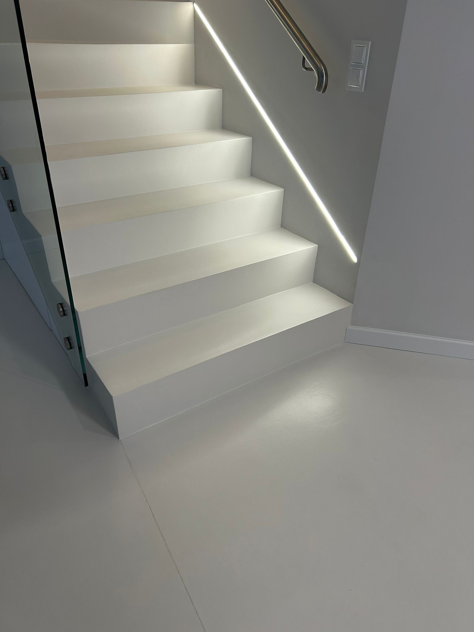 Białe schody z żywicy z podświetleniem LED i szklaną balustradą. Minimalistyczny design, gładka powierzchnia, nowoczesne wykończenie wnętrza.