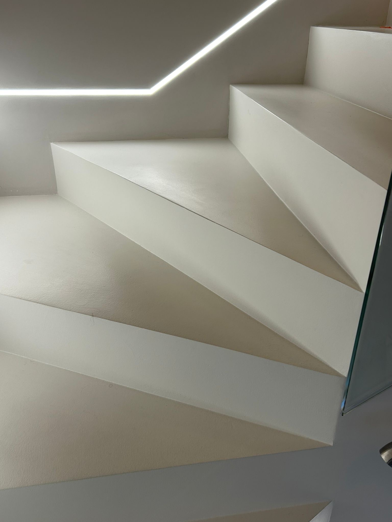 Minimalistyczne schody z mikrocementu w Warszawie, oświetlone listwą LED. Nowoczesne wykończenie wnętrz, jasna kolorystyka, szklana balustrada.