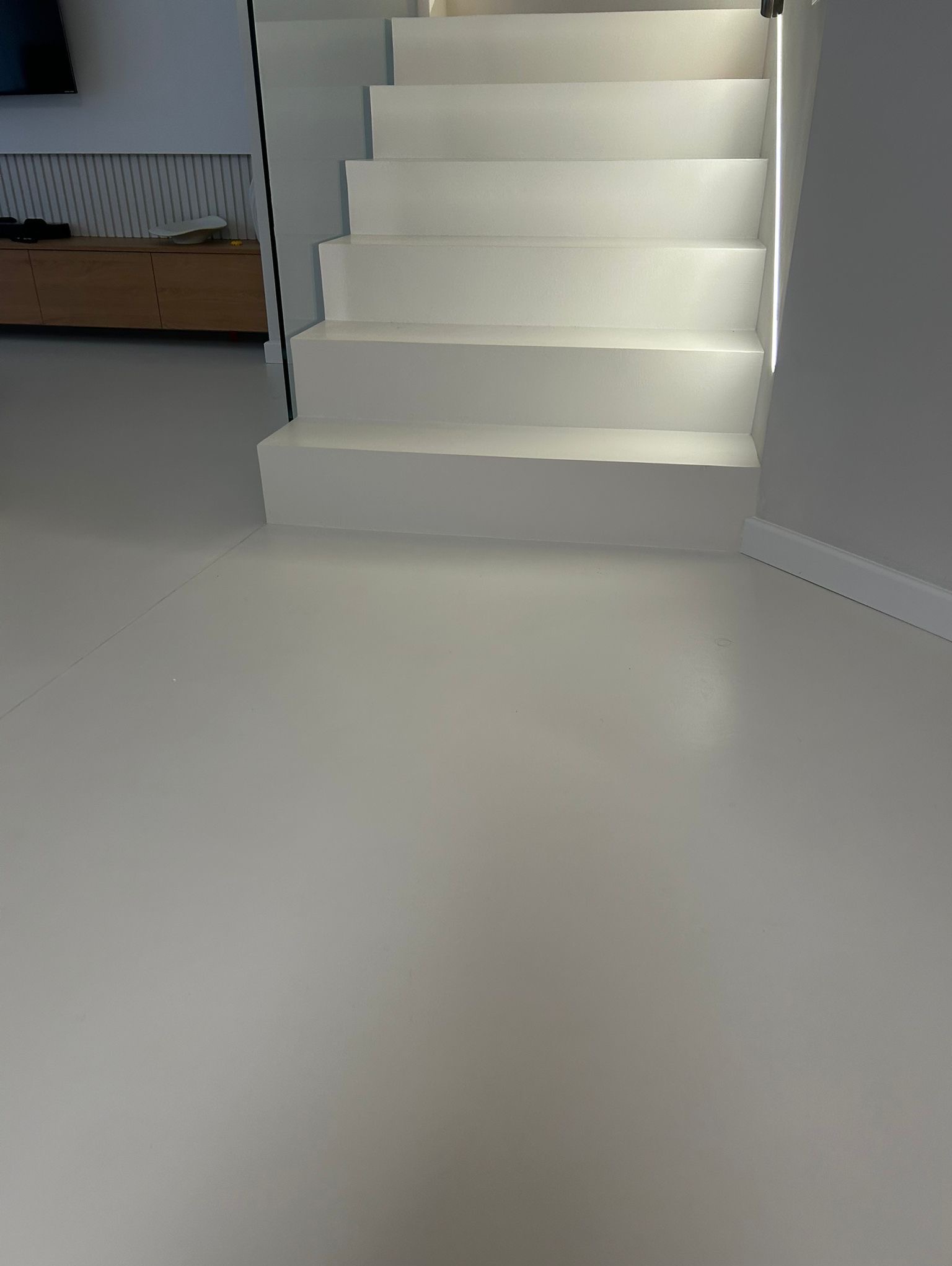 Minimalistyczne, białe schody z gładką, jednolitą posadzką w odcieniach szarości. Widoczny fragment salonu z drewnianą komodą. Nowoczesne wykończenie.