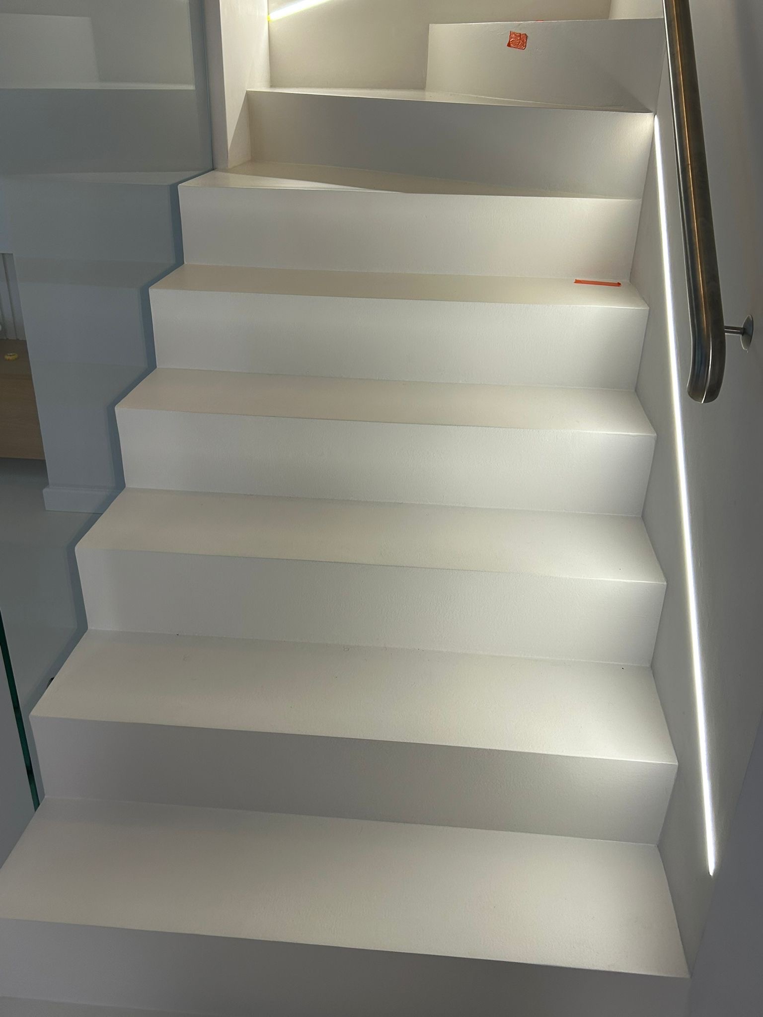 Białe schody z oświetleniem LED wzdłuż ściany i metalową poręczą. Minimalistyczny design, gładka powierzchnia stopni, nowoczesne wykończenie wnętrza.