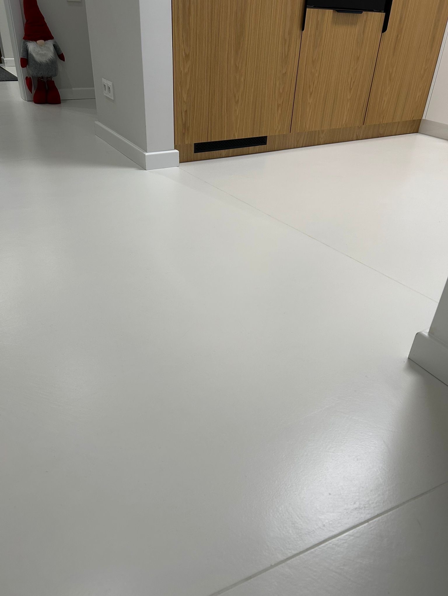 Jasna, jednolita posadzka w nowoczesnym wnętrzu z drewnianą zabudową i dekoracyjnym skrzatem w rogu. Minimalistyczny design, gładka powierzchnia.