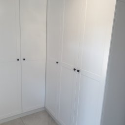 Biała szafa wnękowa z ciemnymi, okrągłymi uchwytami, dopasowana do rogu pomieszczenia. Minimalistyczny design, jasne wnętrze, widoczne płytki podłogowe.