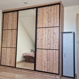 Trzydrzwiowa szafa przesuwna z lustrem i drewnopodobnymi frontami, czarne obramowania, widoczny fragment pokoju z łóżkiem, podłoga z paneli.