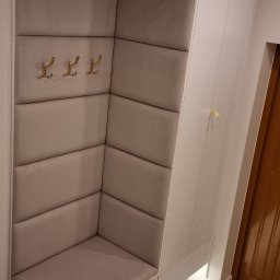 Tapicerowany narożnik z siedziskiem i wieszakami w przedpokoju, uzupełniony białą szafą. Całość oświetlona od dołu, na tle marmurowej podłogi.