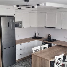 PIJAR Daniel Przesmycki - Nowoczesna kuchnia w bieli i drewnie: zabudowa z wyspą, czarne akcenty, oświetlenie sufitowe. Widok z góry na gotową realizację.