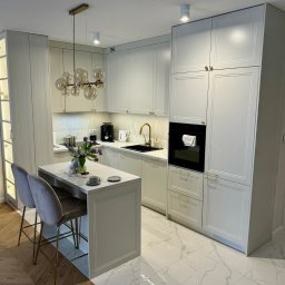 PIJAR Daniel Przesmycki - Elegancka, jasna kuchnia z wyspą i złotymi akcentami: bateria, uchwyty, lampa. Marmurowe blaty i podłoga. Nowoczesny design z kwiatami i kawą na blacie.