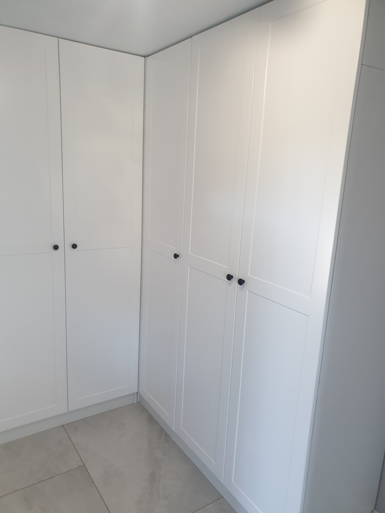 Biała szafa wnękowa z ciemnymi, okrągłymi uchwytami, dopasowana do rogu pomieszczenia. Minimalistyczny design, jasne wnętrze, widoczne płytki podłogowe.