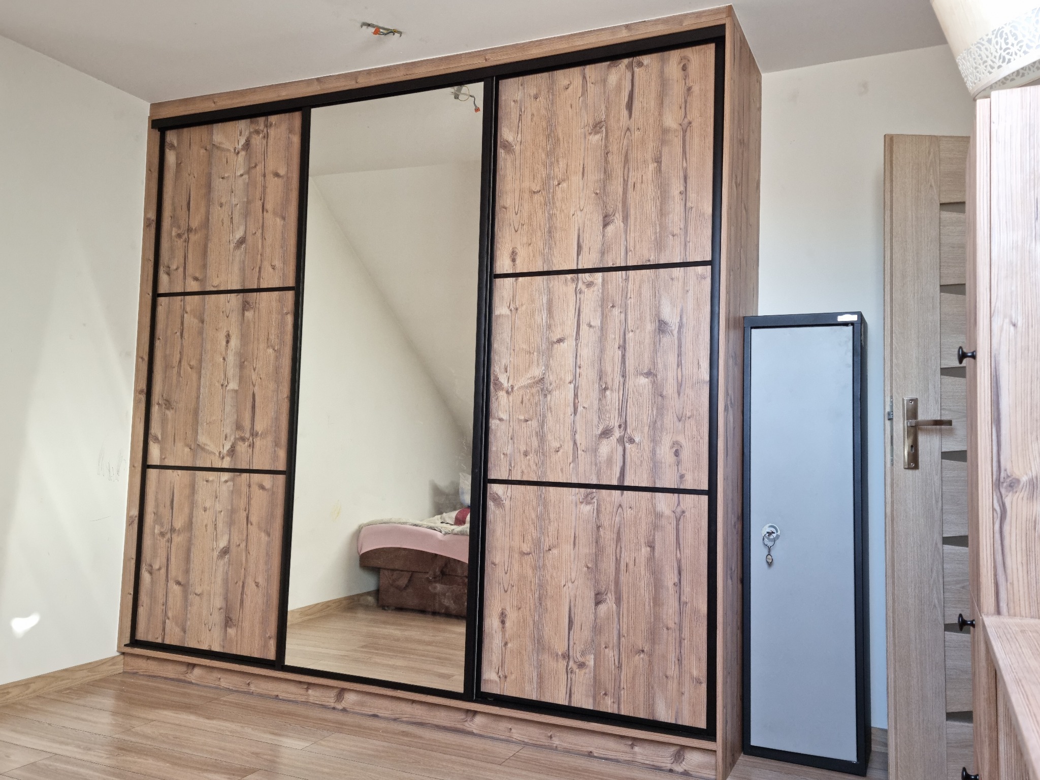 Trzydrzwiowa szafa przesuwna z lustrem i drewnopodobnymi frontami, czarne obramowania, widoczny fragment pokoju z łóżkiem, podłoga z paneli.