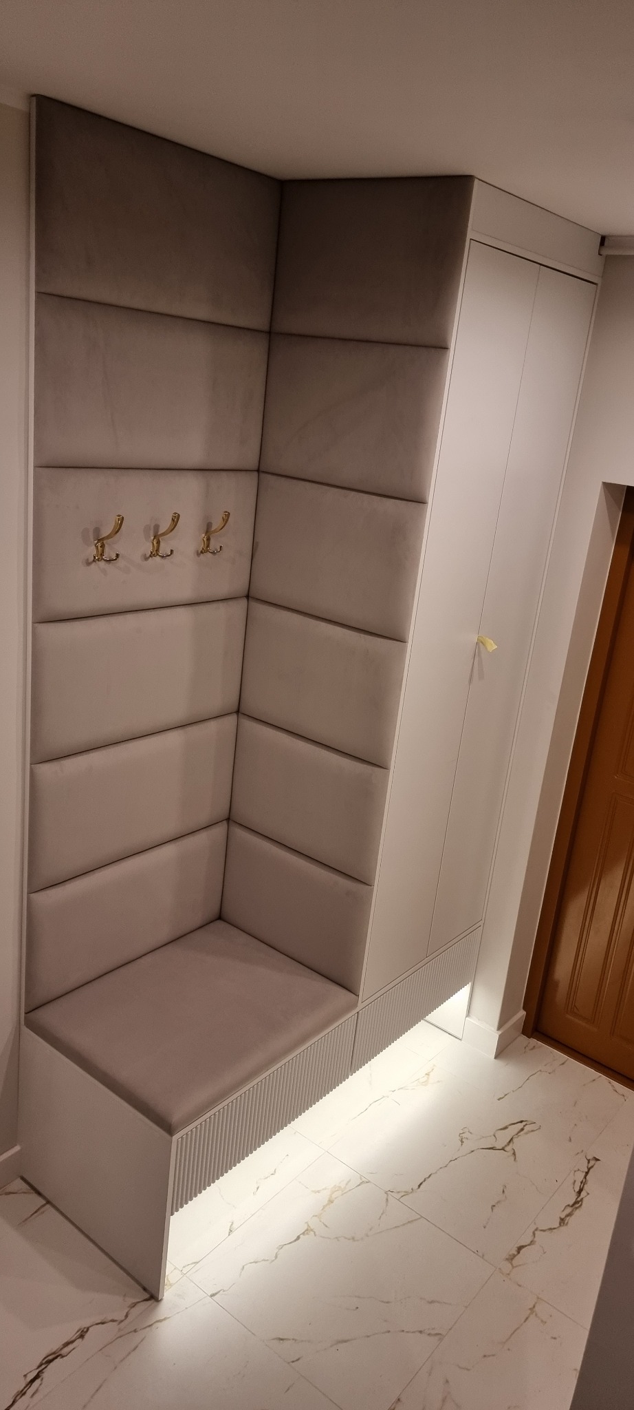 Tapicerowany narożnik z siedziskiem i wieszakami w przedpokoju, uzupełniony białą szafą. Całość oświetlona od dołu, na tle marmurowej podłogi.