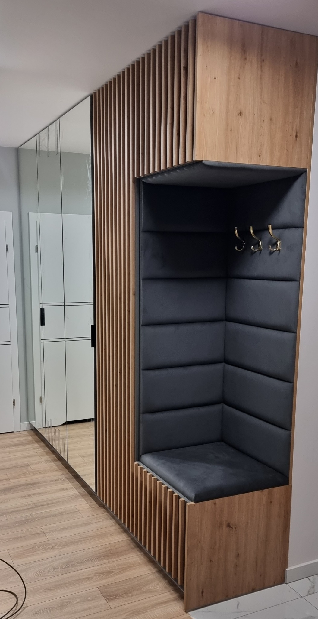 Nowoczesna zabudowa przedpokoju z siedziskiem tapicerowanym, wnęką na wieszaki i lustrem. Drewniane lamele tworzą ażurową ściankę. Minimalistyczny design.