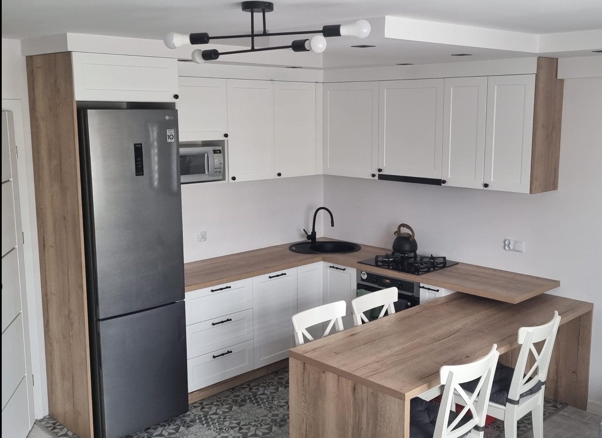 Nowoczesna kuchnia w bieli i drewnie: zabudowa z wyspą, czarne akcenty, oświetlenie sufitowe. Widok z góry na gotową realizację.