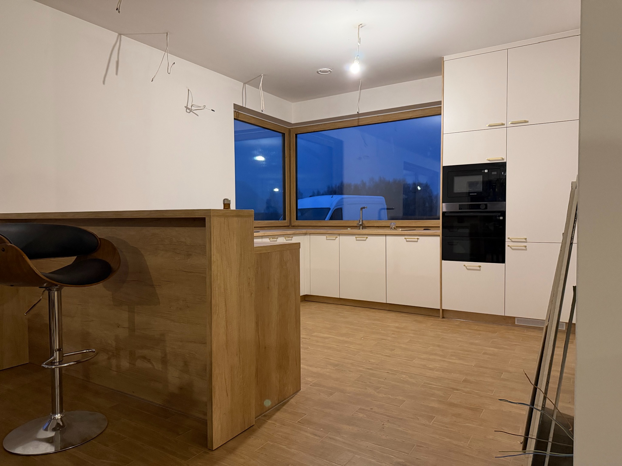 Jasna kuchnia z drewnianym blatem i oknem z widokiem na zewnątrz. Hoker przy blacie. Częściowo widoczne kable elektryczne na ścianie, sugerujące prace wykończeniowe.