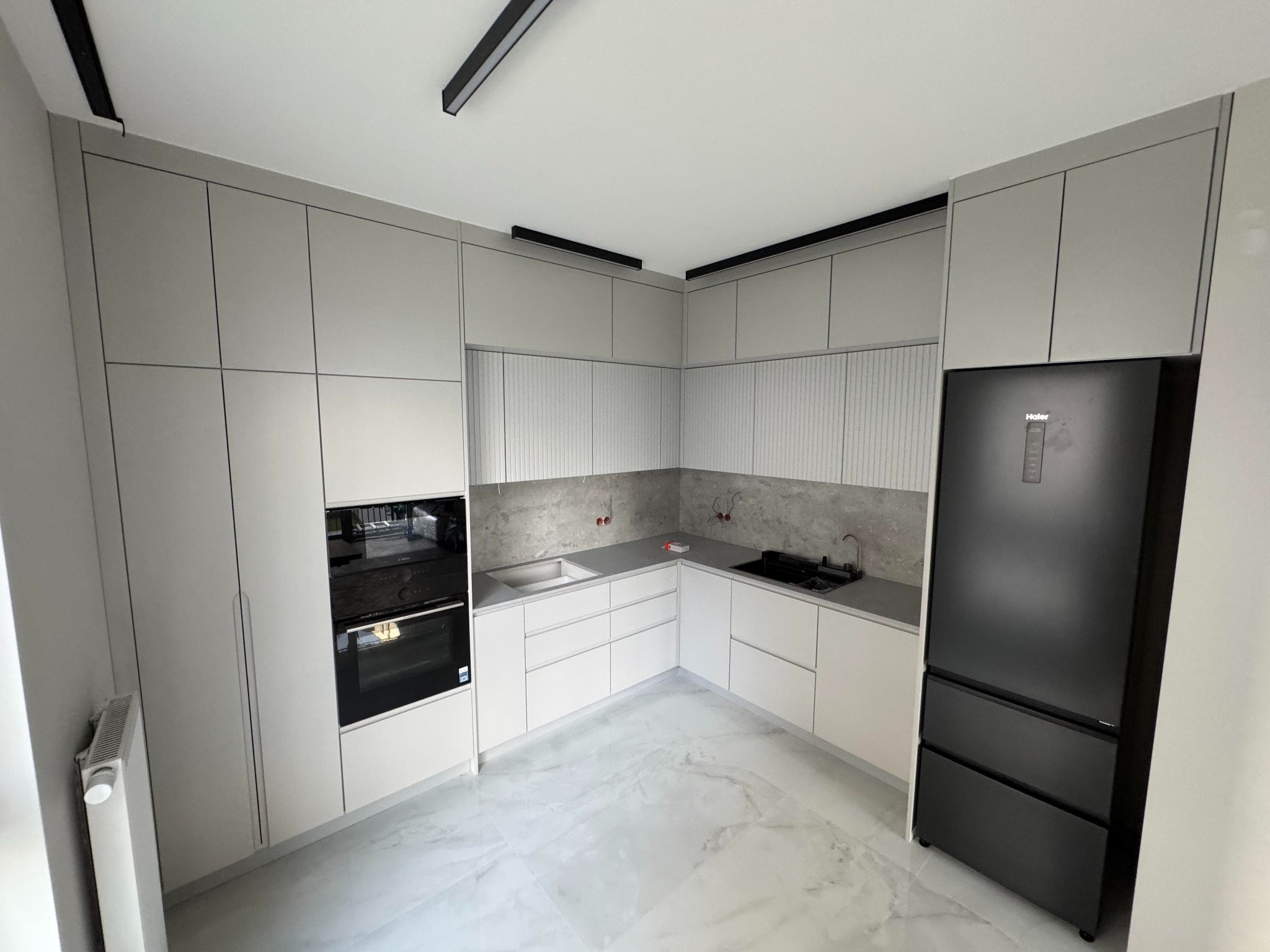 Jasna kuchnia z szarymi frontami, czarnym sprzętem AGD i oświetleniem szynowym. Minimalistyczny design, marmurowa podłoga, widok na całe pomieszczenie.