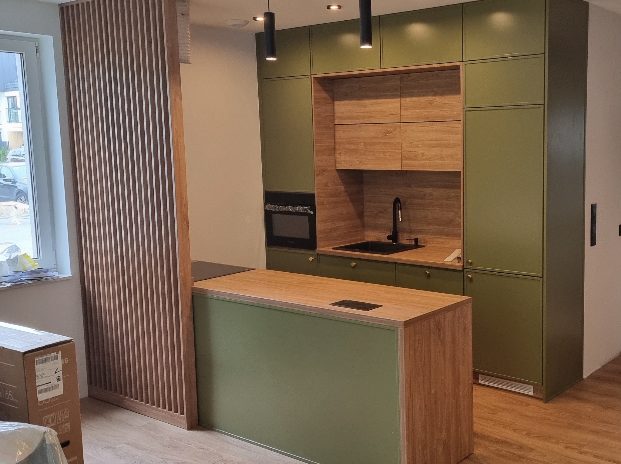 Nowoczesna kuchnia w odcieniach zieleni i drewna z wyspą, oświetleniem punktowym i dekoracyjną ścianką z drewnianych listew. Minimalistyczny design.