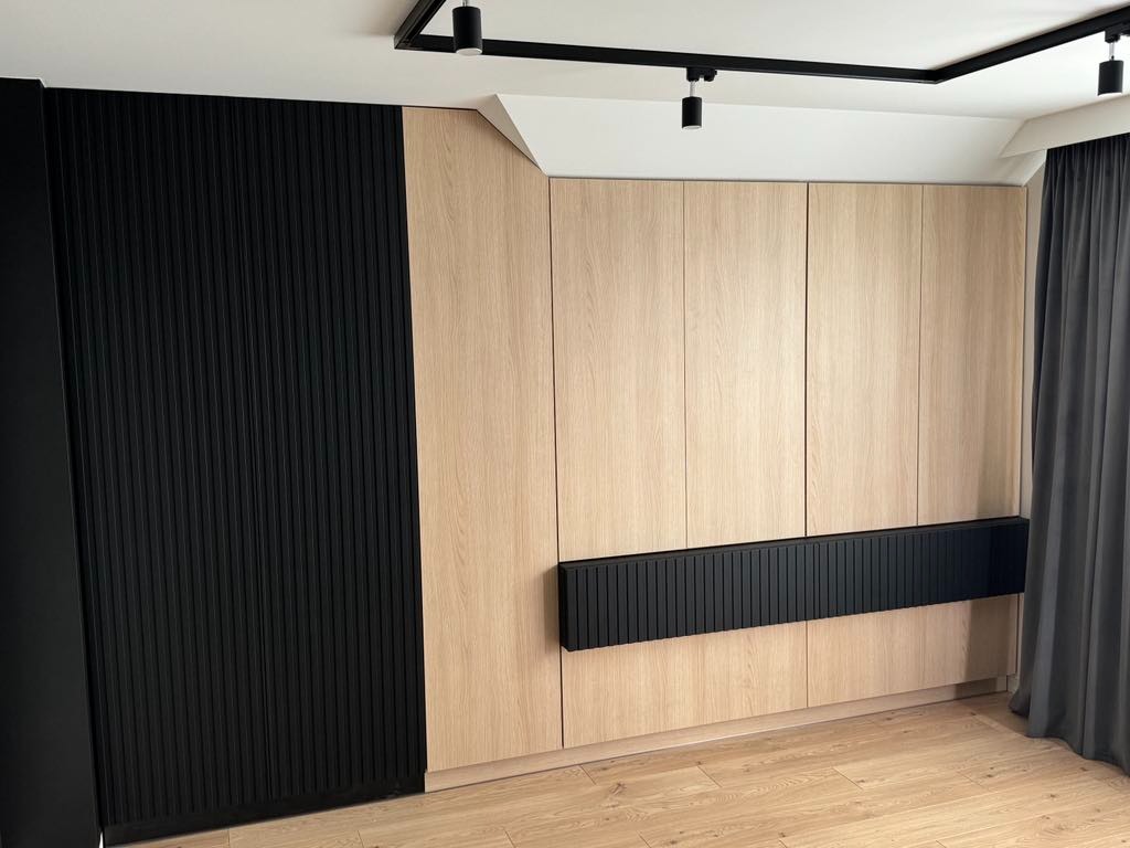 Nowoczesna szafa wnękowa z jasnego drewna i czarnymi, ryflowanymi akcentami. Minimalistyczny design, oświetlenie punktowe, drewniana podłoga i szare zasłony w tle.