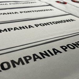 HEKATE Personalizacja odzieży kraśnik - Białe koszulki z czarnym nadrukiem 'KOMPANIA PONTONOWA' ułożone rzędami, widoczne szwy i faktura materiału. Perspektywa z góry, ostre światło.