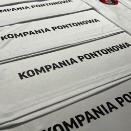 HEKATE Personalizacja odzieży kraśnik - Stos białych koszulek z czarnym nadrukiem 'KOMPANIA PONTONOWA' i logo. Ułożone równo, widoczne szwy i detale materiału. Perspektywa z góry, oświetlenie studyjne.