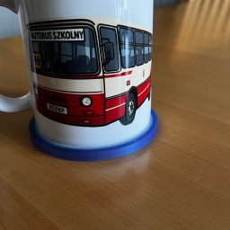 HEKATE Personalizacja odzieży kraśnik - Ceramiczny kubek z nadrukiem autobusu szkolnego, stojący na niebieskiej podkładce na drewnianym stole. Kubek ma czerwone obramowanie i napis 'Autobus Szkolny' oraz imię 'Józef'.